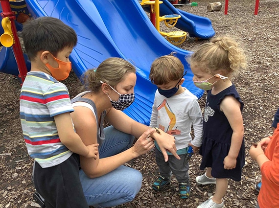 3 young children and a teacher stand in front of blue slides on a playground