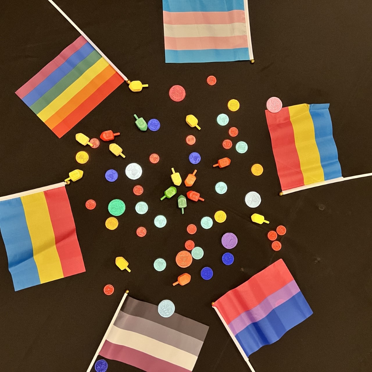 Rainbow pride flags on a table surrounding colored dreidels and gelt