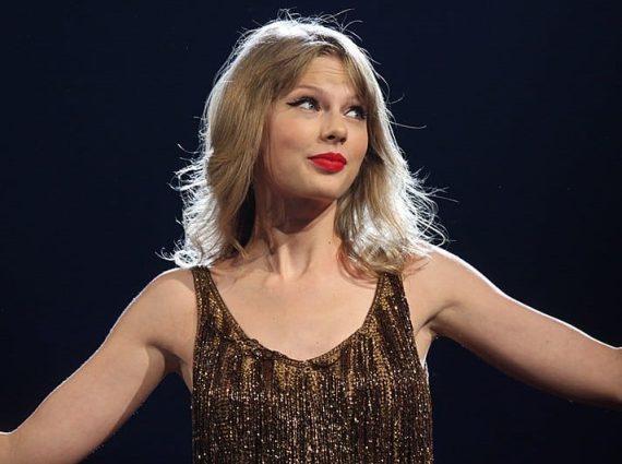 Taylor Swift standing with outstretched arms with a black background behind her