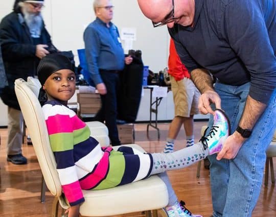 Young Black girl in colorful striped sweater sits in a chair while a volunteer helps her put boots on