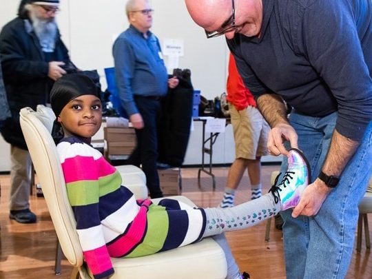 Young Black girl in colorful striped sweater sits in a chair while a volunteer helps her put boots on