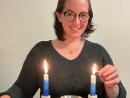 Lizzie Gomes in front of a white wall lighting two blue and white Shabbat candles.
