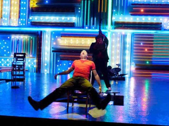 actor in red shirt sits onstage with bright colored lights behind him