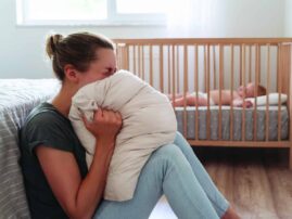 Woman sits on floor in front of crib, screaming into a pillow