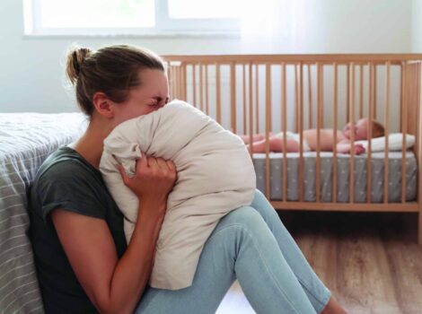 Woman sits on floor in front of crib, screaming into a pillow