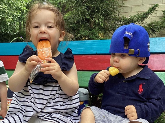 Two young children sit on a bench eating popsicles