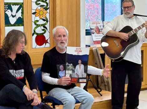 Two people sit in chairs holding a photo of an Israeli hostage while a man playing guitar stands next to them