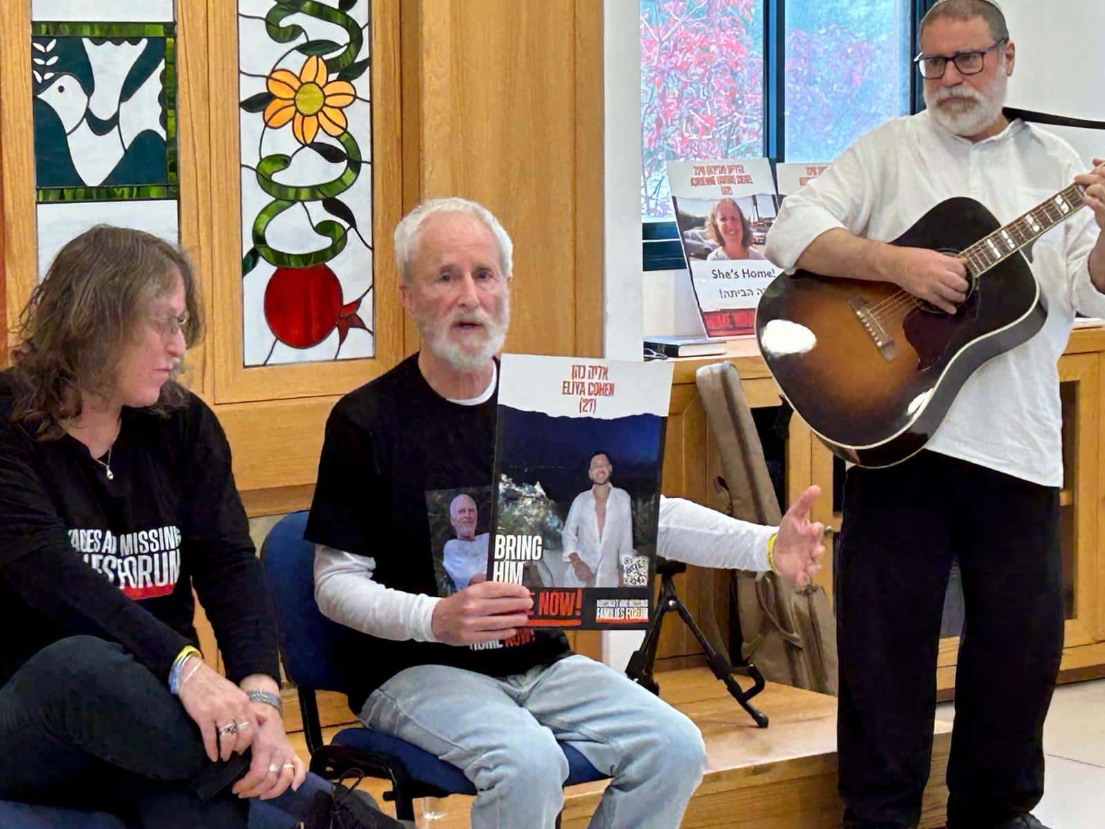 Two people sit in chairs holding a photo of an Israeli hostage while a man playing guitar stands next to them