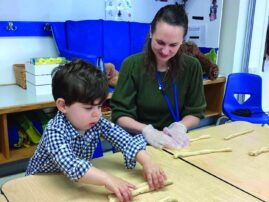 young boy rolls out challah dough while Kristin Zeldes observes