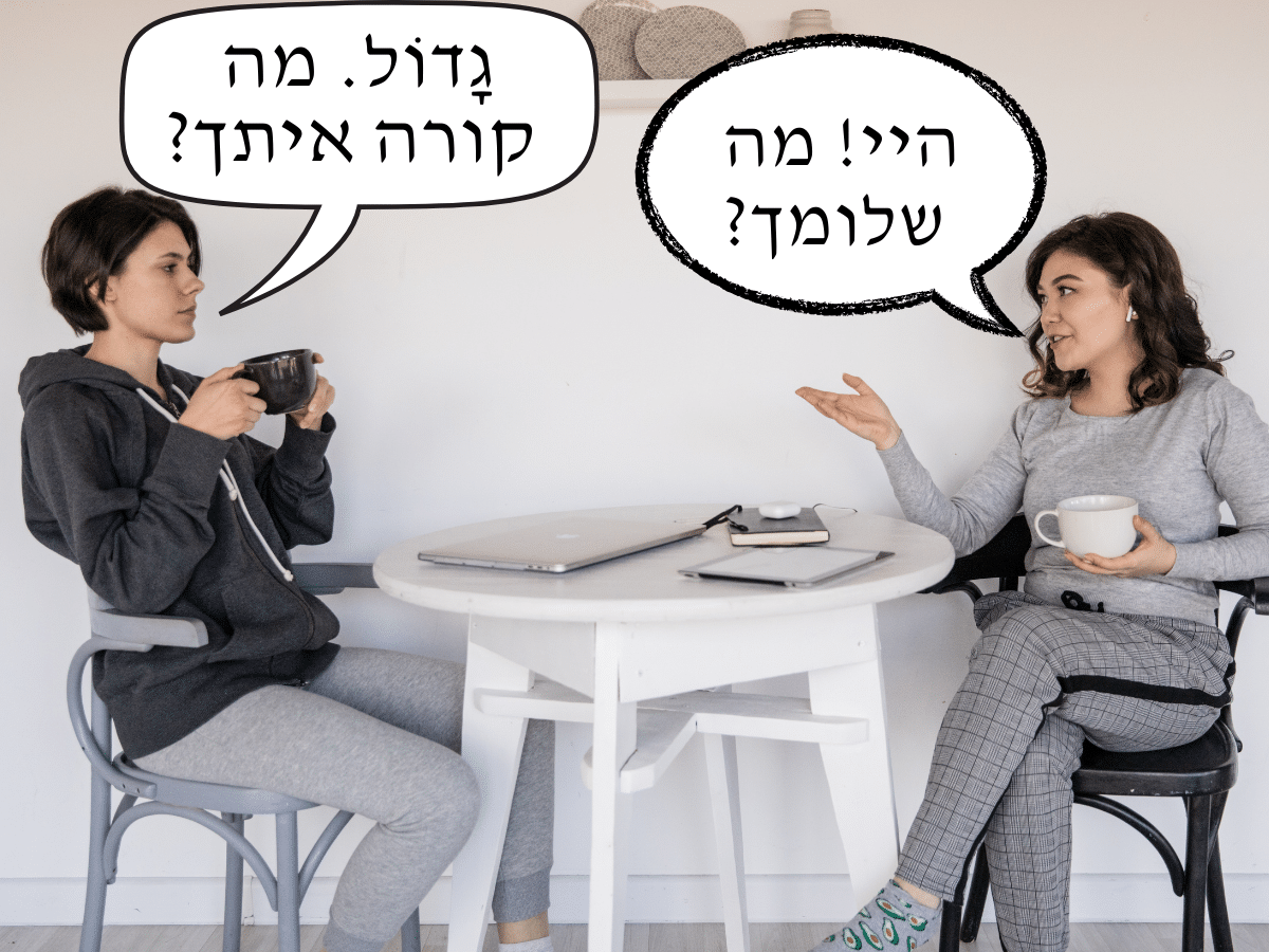 Two women sit at a table chatting, with conversation bubbles above them with Hebrew text.