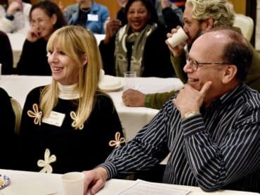 Adult man and woman sit at a table listening to a facilitator during the 12 Jewish Questions Class