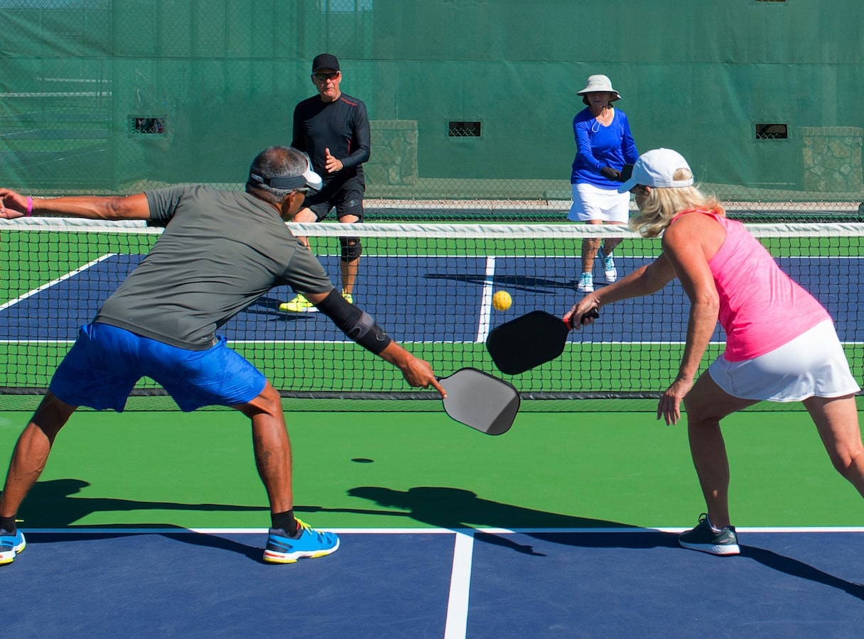A man and a woman reach their pickleball paddles toward the ball while another pair waits to receive it.